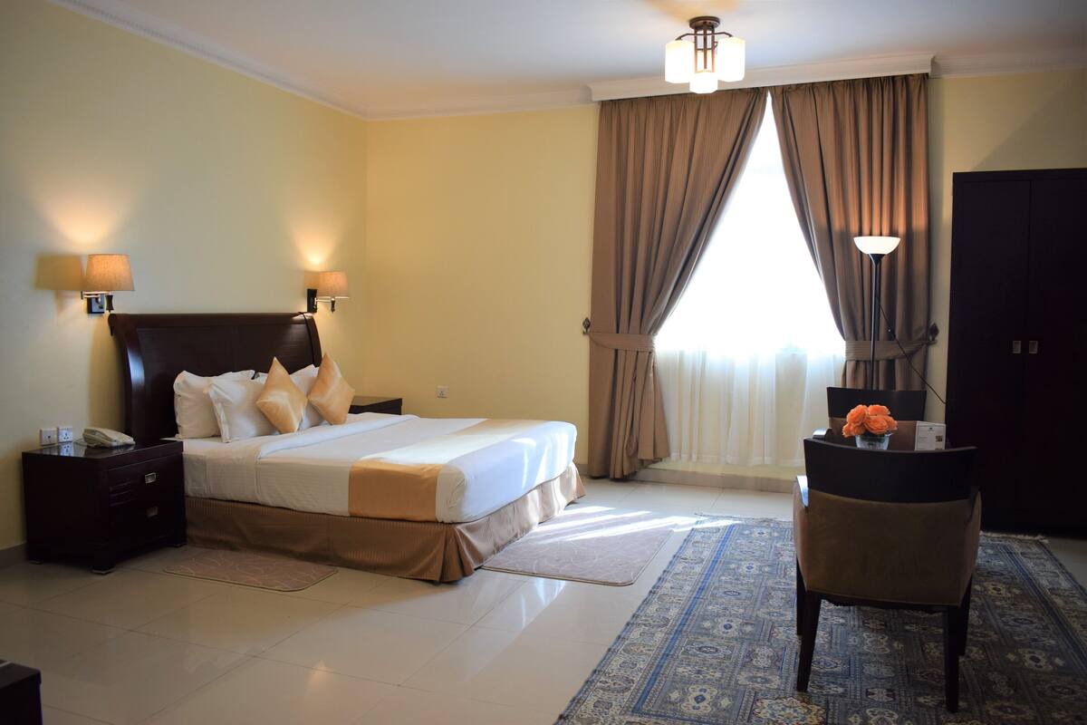 Una habitación de hotel con cama, mesitas de noche, un escritorio con silla, lámpara y ventana con cortinas.