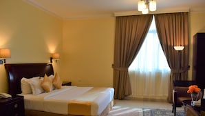 A hotel room with a large bed, two bedside lamps, a desk with a chair, and a view of the outdoors through a window with curtains. - Crown Palace Hotel Ajman (Ajman)