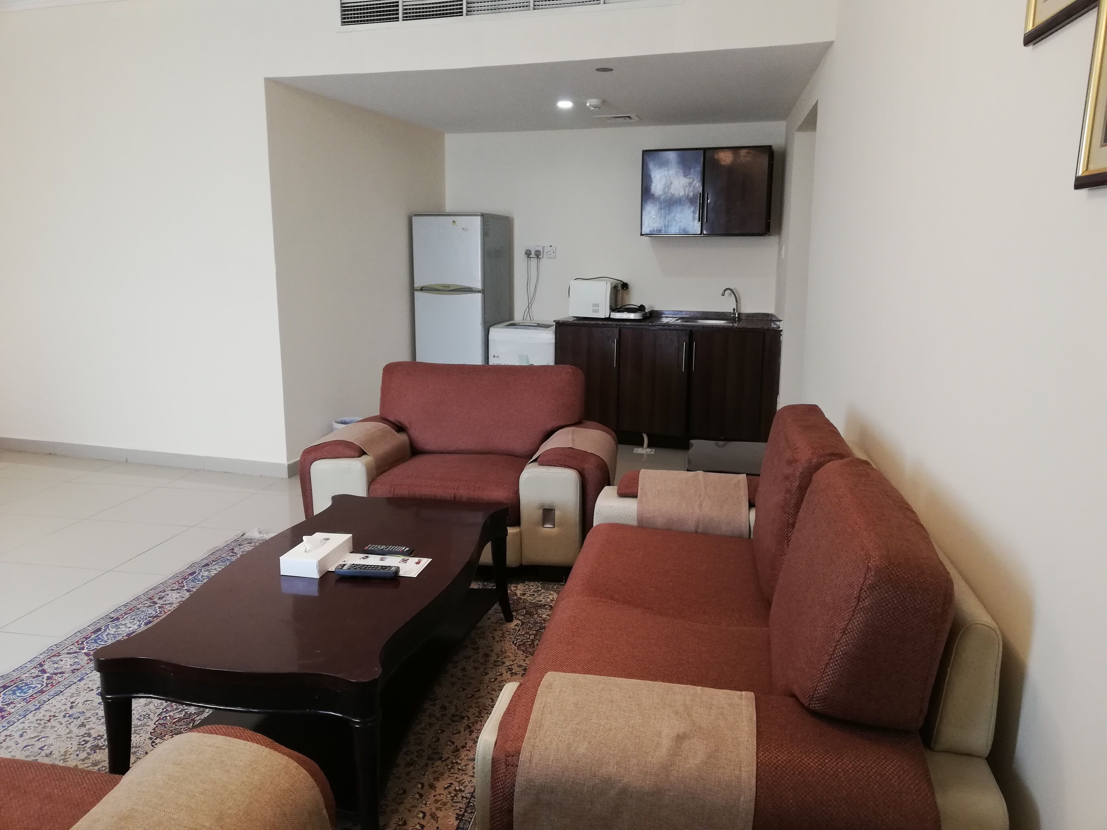 A living room with a sofa, armchairs, and a coffee table. There is a kitchen area with a refrigerator, sink, and microwave in the background.