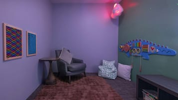 Una habitación con pared morada, decoración de dinosaurio azul en la pared, un sillón gris, una mesita redonda pequeña y una alfombra marrón y esponjosa.