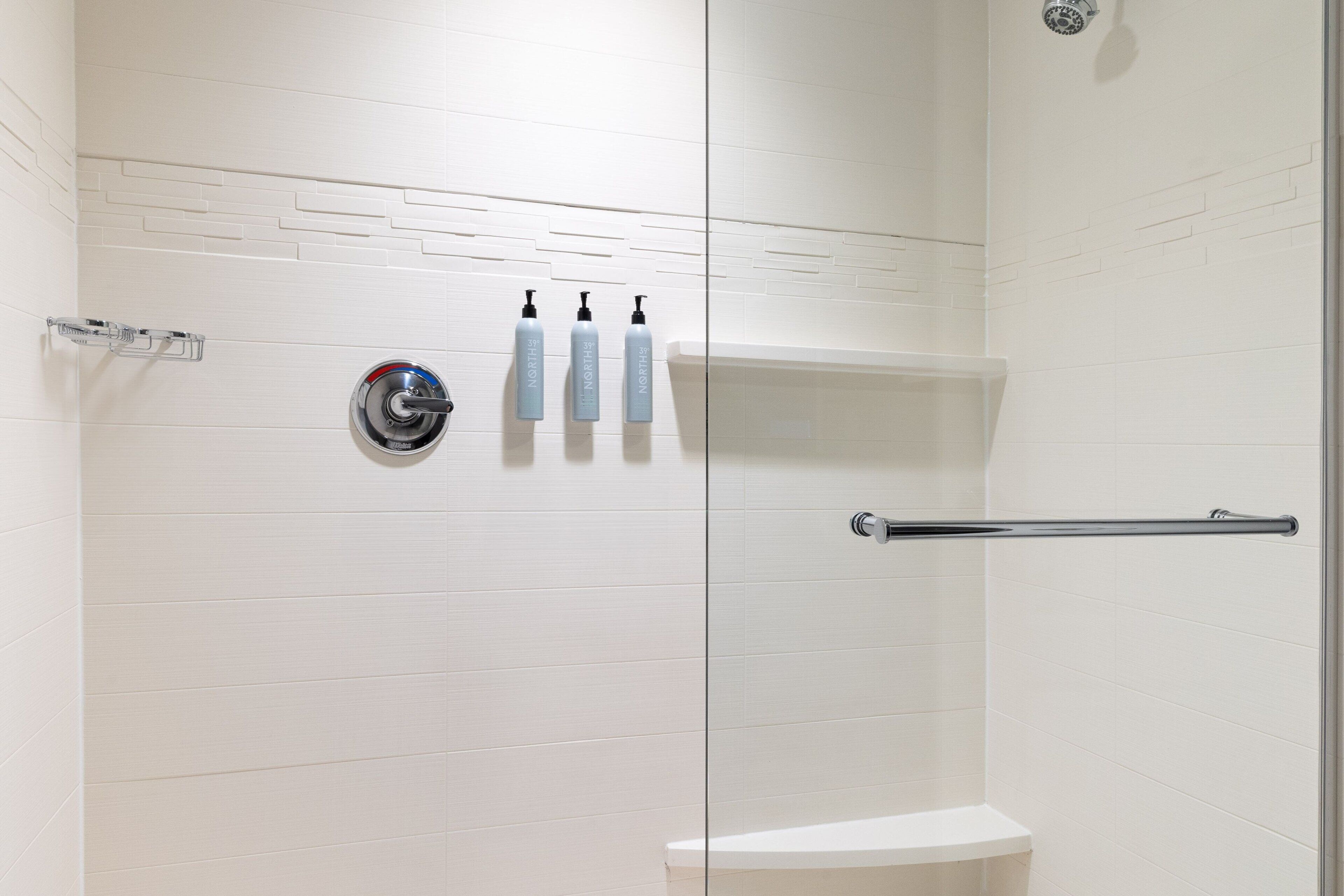 A modern bathroom with white tiled walls, a glass shower enclosure, a toilet, and three bottles of hand soap on the wall.