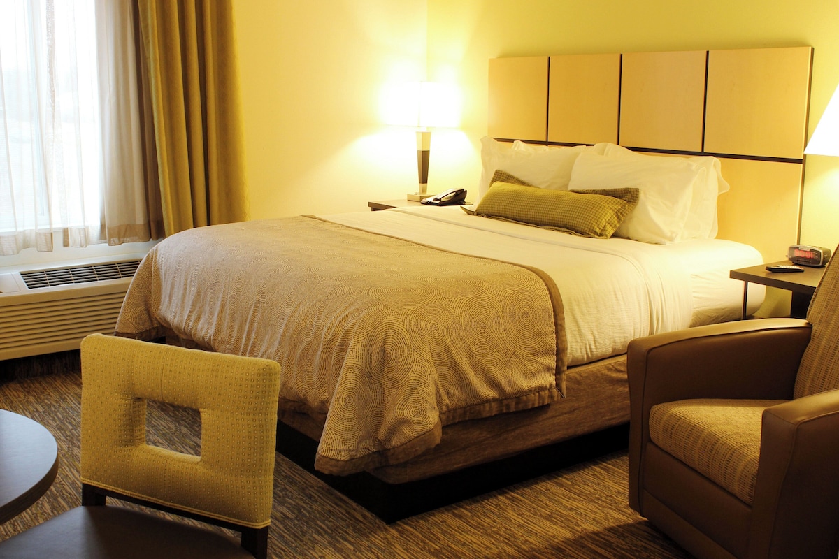 Quarto de hotel com cama, duas luminárias de cabeceira, uma cadeira, uma mesinha e uma janela com cortinas.