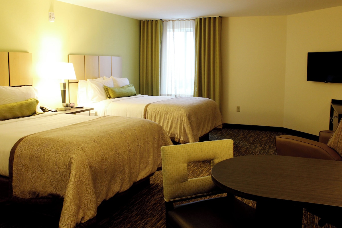 Quarto de hotel com duas camas, uma escrivaninha, uma cadeira, uma televisão e janela com cortinas.