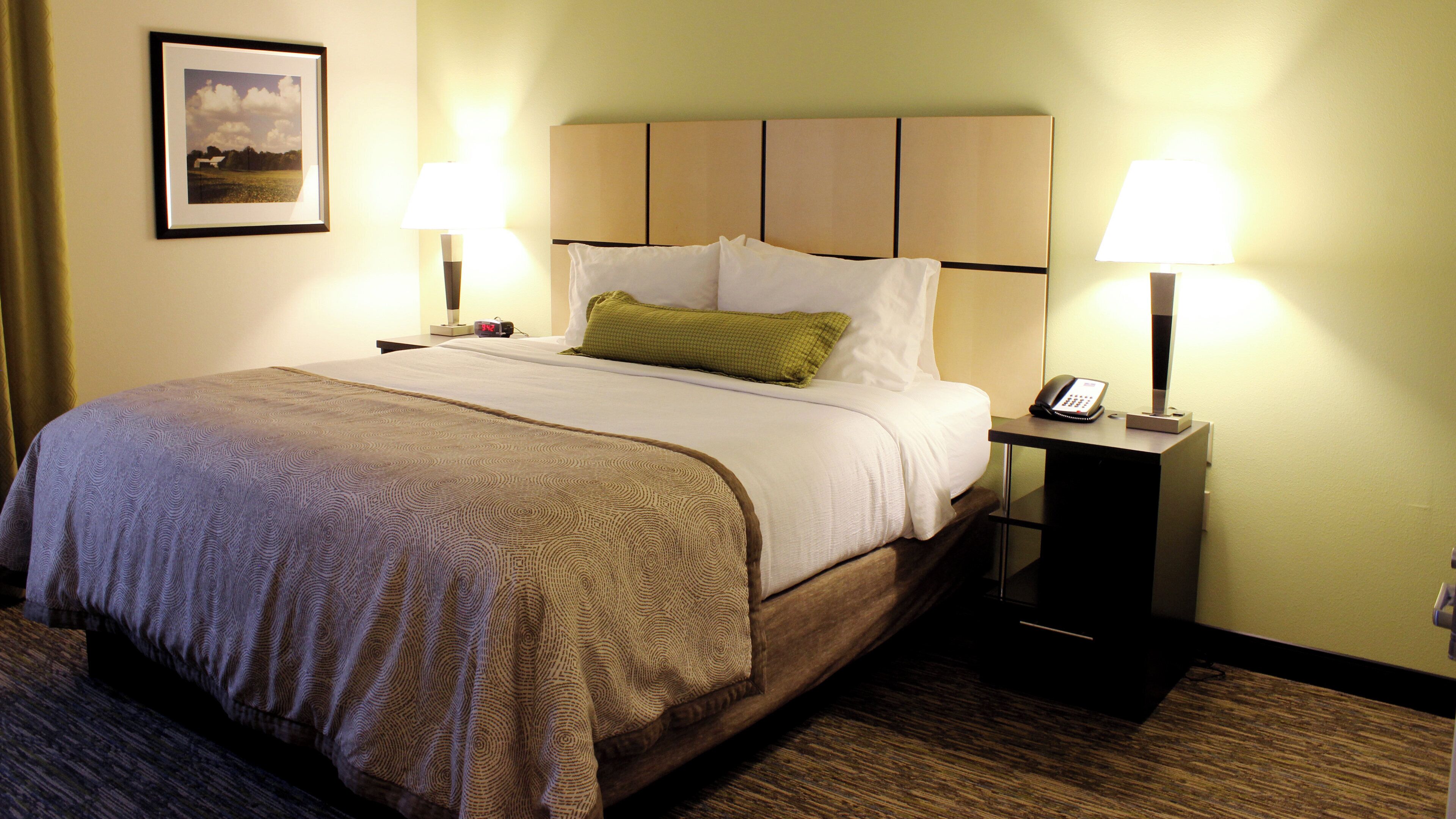 A hotel room with a bed, two bedside lamps, a nightstand with a phone, and a framed picture on the wall.