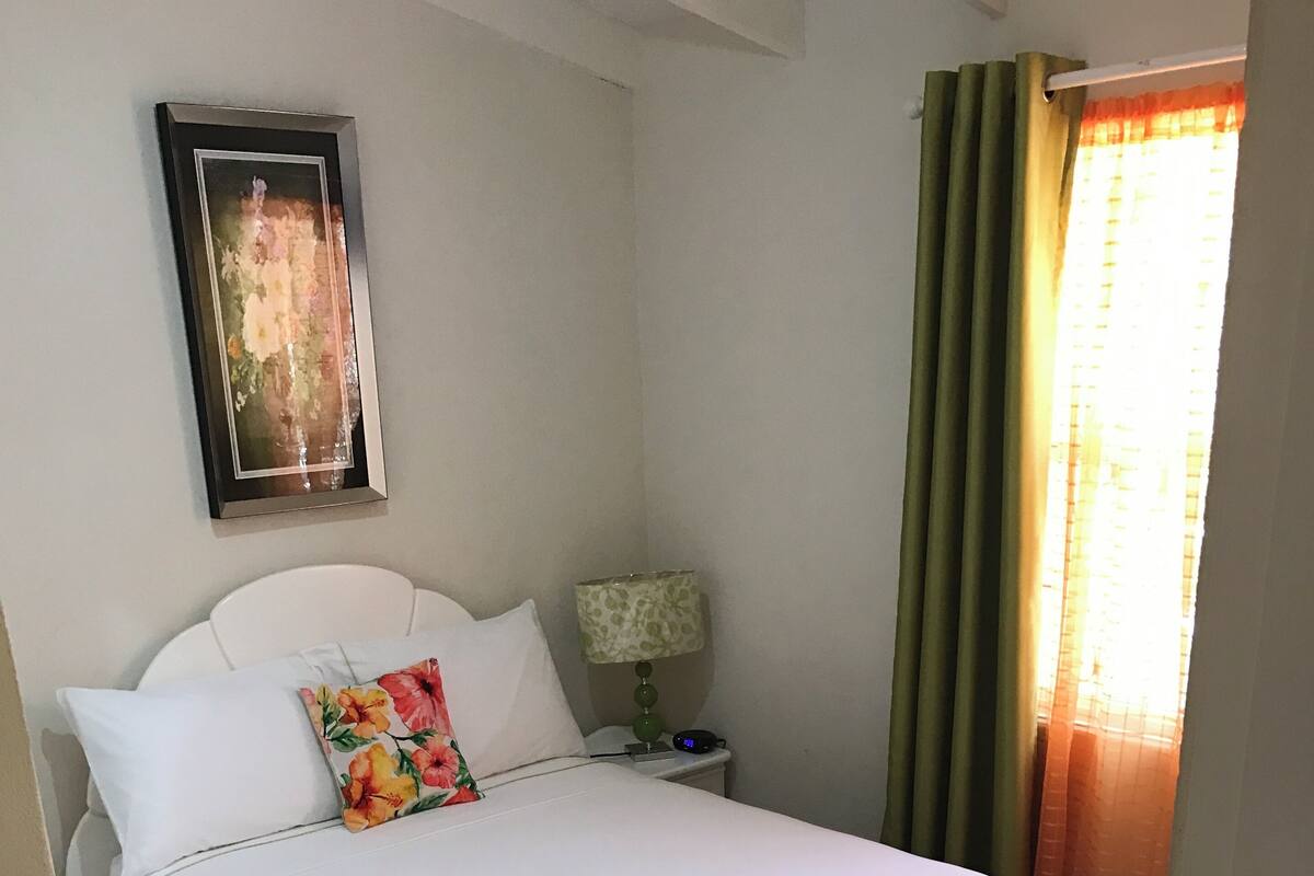 Una cama bien hecha con ropa blanca, una almohada estampada con flores y una mesita de noche con una lámpara y un reloj digital.
