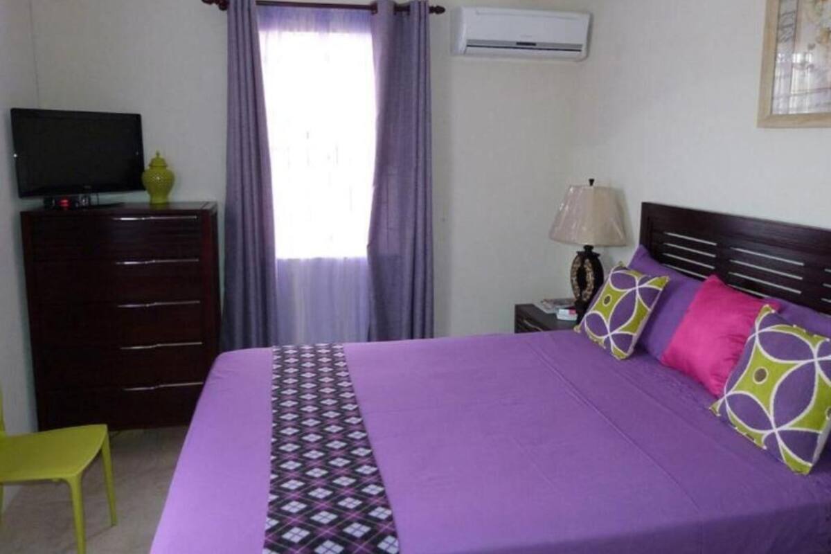 Un dormitorio con cama, televisión, cómoda, lámpara y ventana con cortinas.