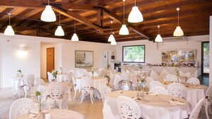 Una stanza spaziosa con tavoli rotondi e sedie bianche, travi di legno al soffitto e un'ampia finestra con vista sul verde.
