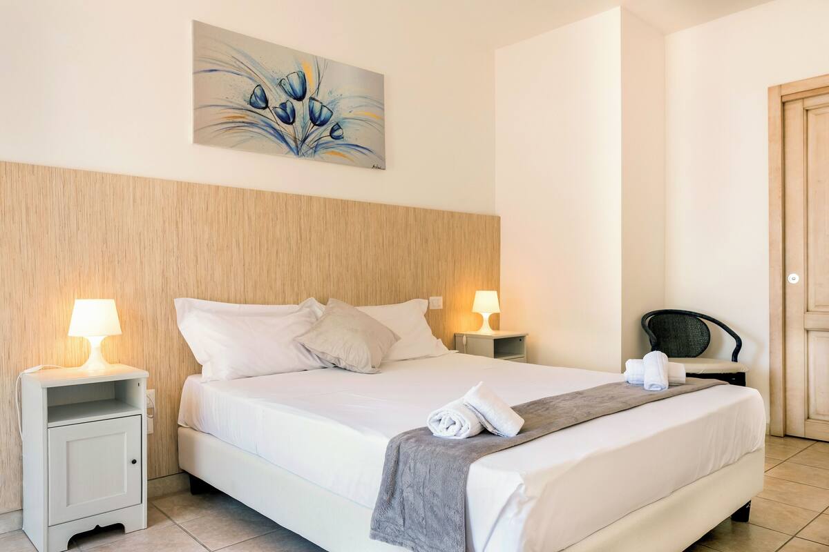Een net opgemaakt bed met witte lakens, twee bedlampjes, een houten hoofdbord, een schilderij boven het bed en een stoel in de hoek.
