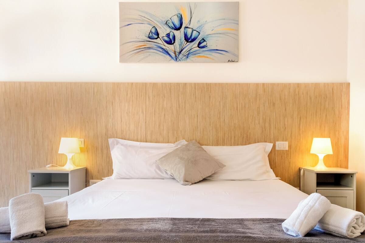 Een netjes opgemaakt bed met witte lakens, twee bedlampjes en een schilderij erboven.
