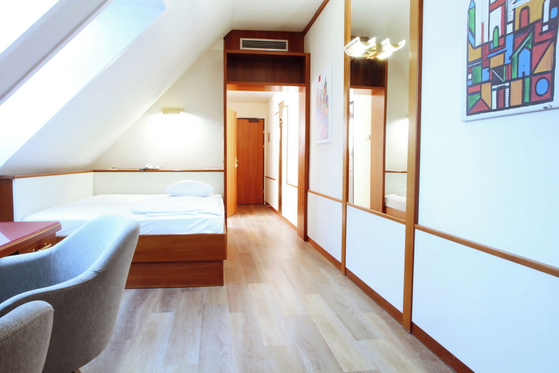 Quarto com teto inclinado, uma cama, uma escrivaninha e uma cadeira. Há um corredor que dá para outro cômodo.