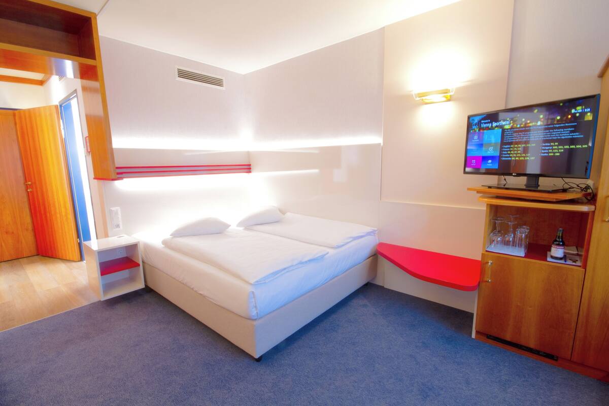 A modern hotel room with a bed, a TV, a wooden cabinet, and a red bench.