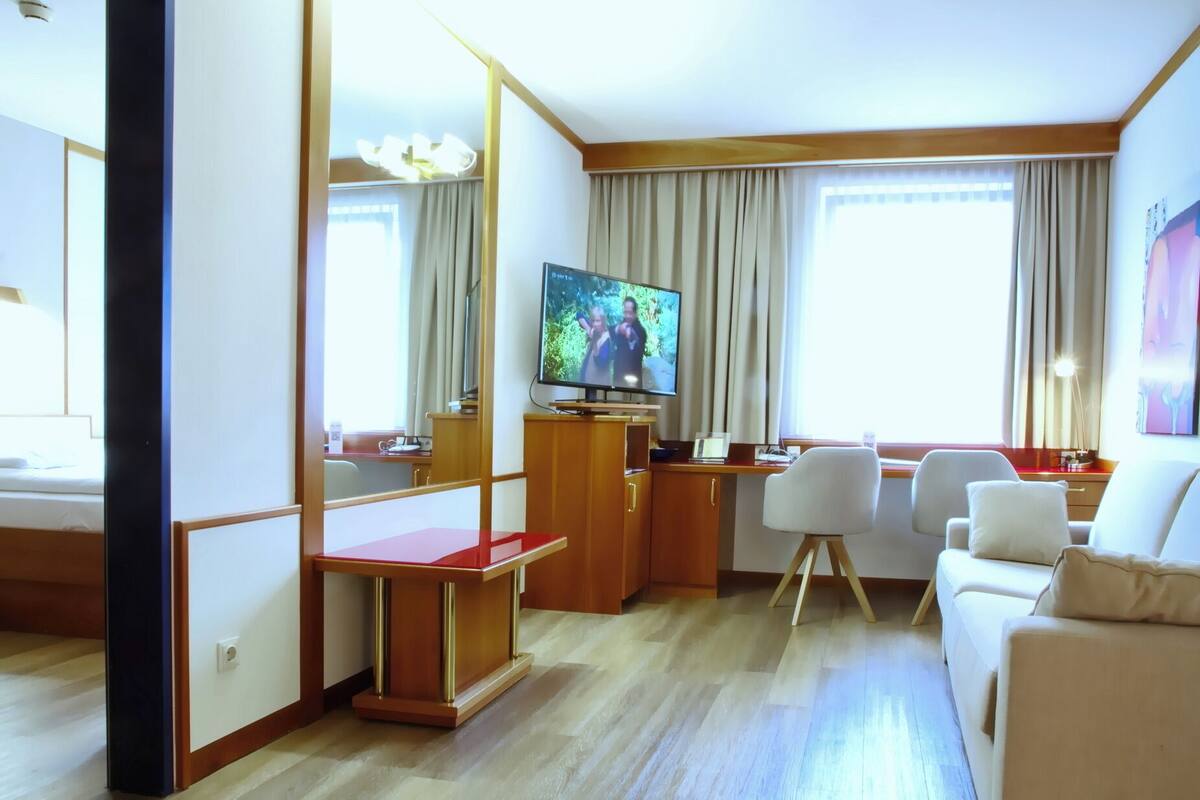 A hotel room with a wooden desk, a television, a sofa, and a large window with curtains.
