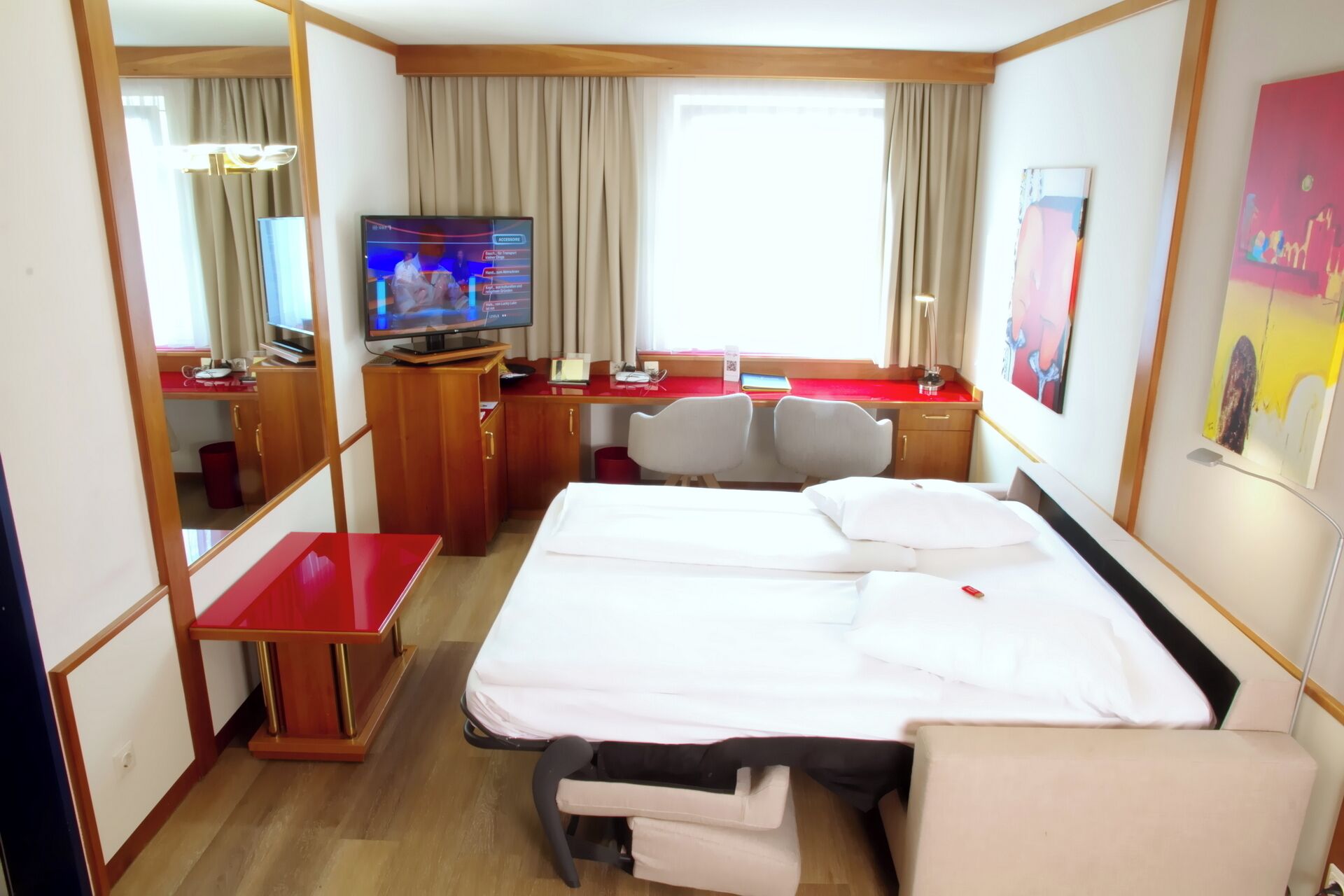 Quarto de hotel com cama, escrivaninha com computador, televisão e janela com cortinas.