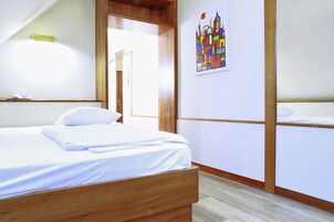 Quarto de hotel com cama de madeira, roupa de cama branca, mesa de cabeceira pequena, luminária na parede e pintura colorida de paisagem urbana.