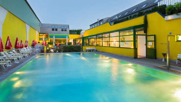 Una piscina con tumbonas y sombrillas, rodeada por un edificio amarillo con ventanas amplias y techo verde.