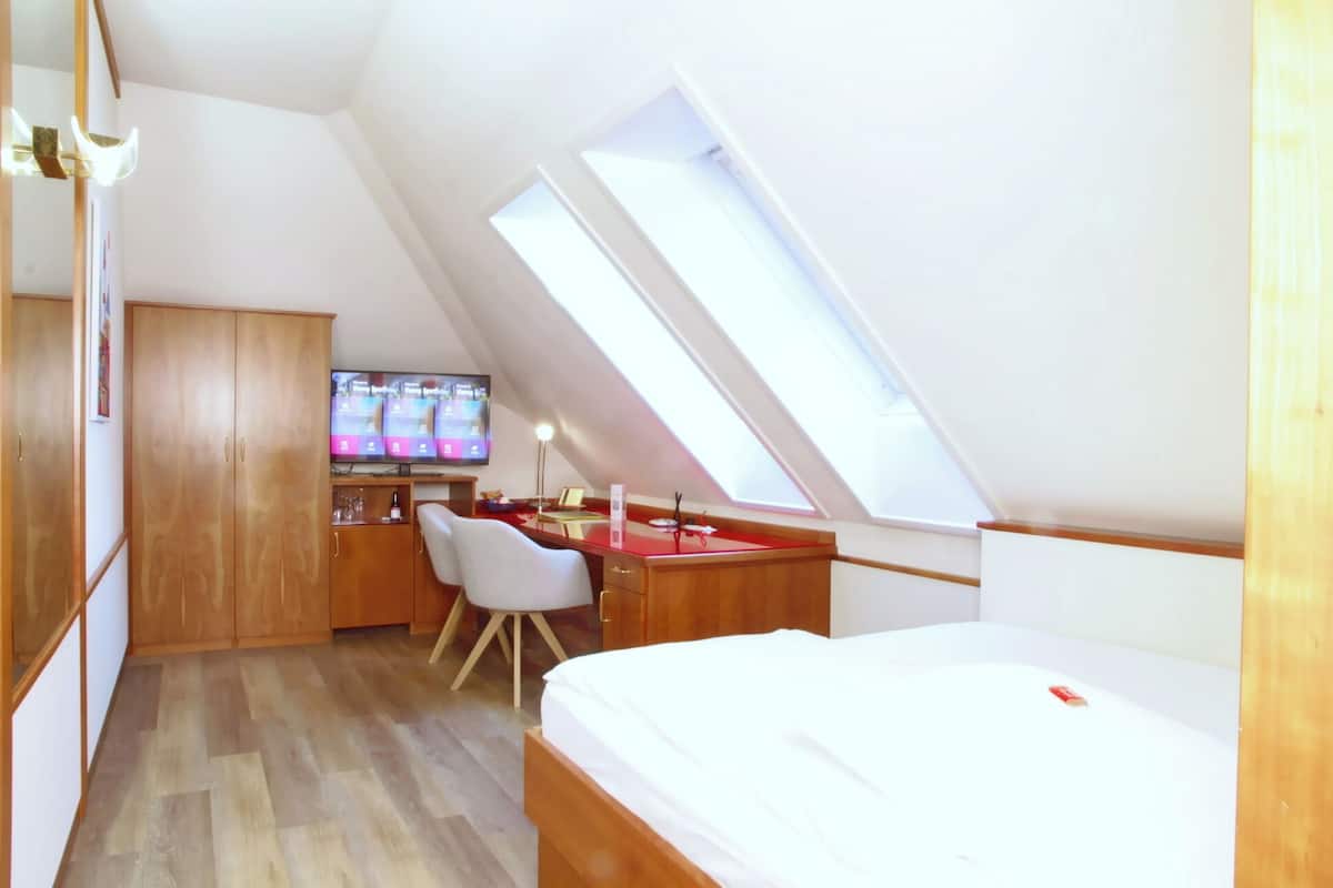 A compact room with a sloped ceiling, a bed, a desk with a chair, and a television.