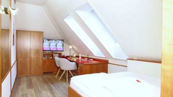 A compact room with a sloped ceiling, a bed, a desk with a chair, and a television.
