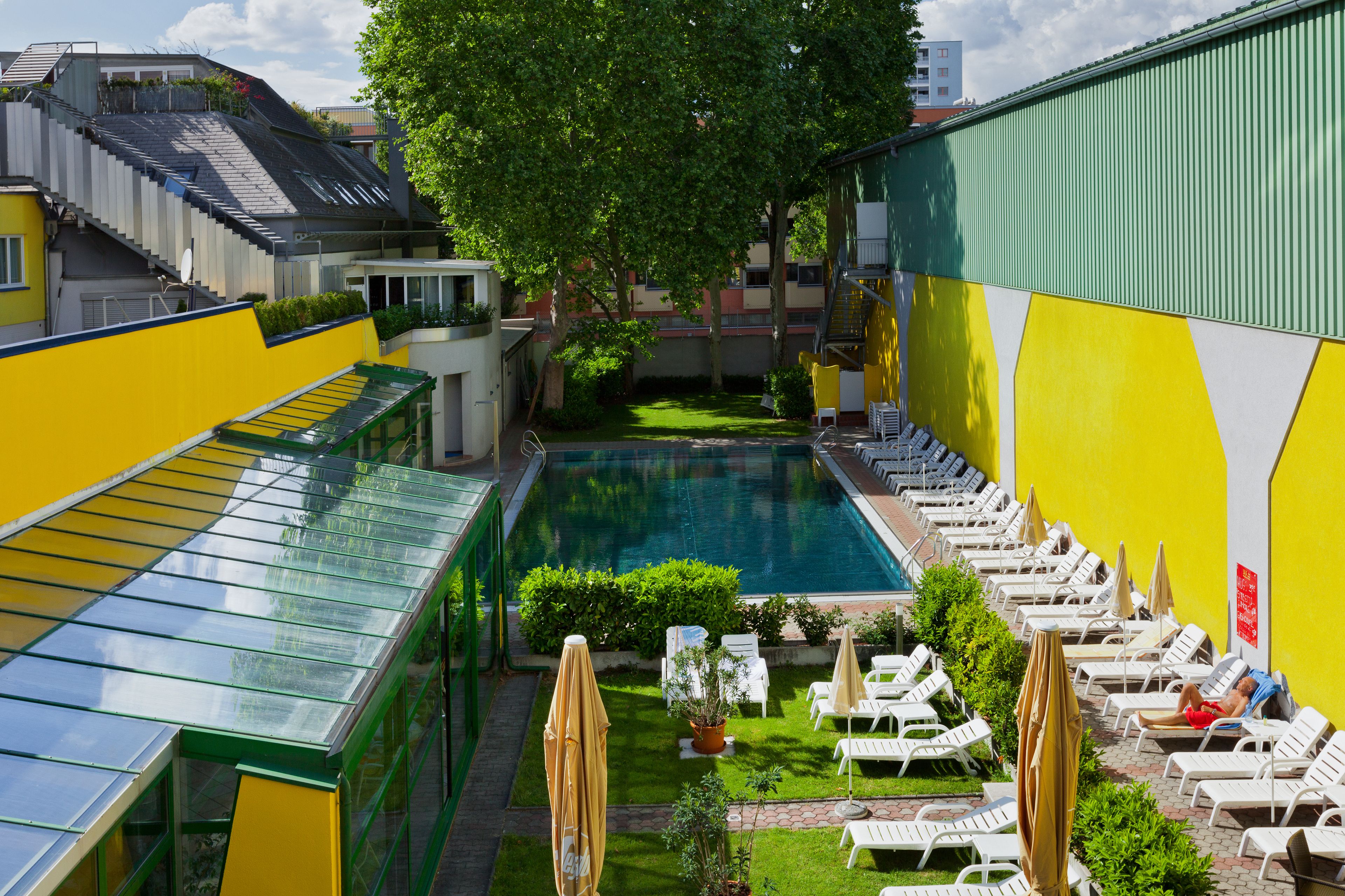 Uma piscina rodeada por espreguiçadeiras e guarda-sóis, com um edifício verde e amarelo ao fundo.
