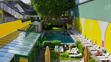 A swimming pool surrounded by sun loungers and umbrellas, with a green and yellow building in the background.