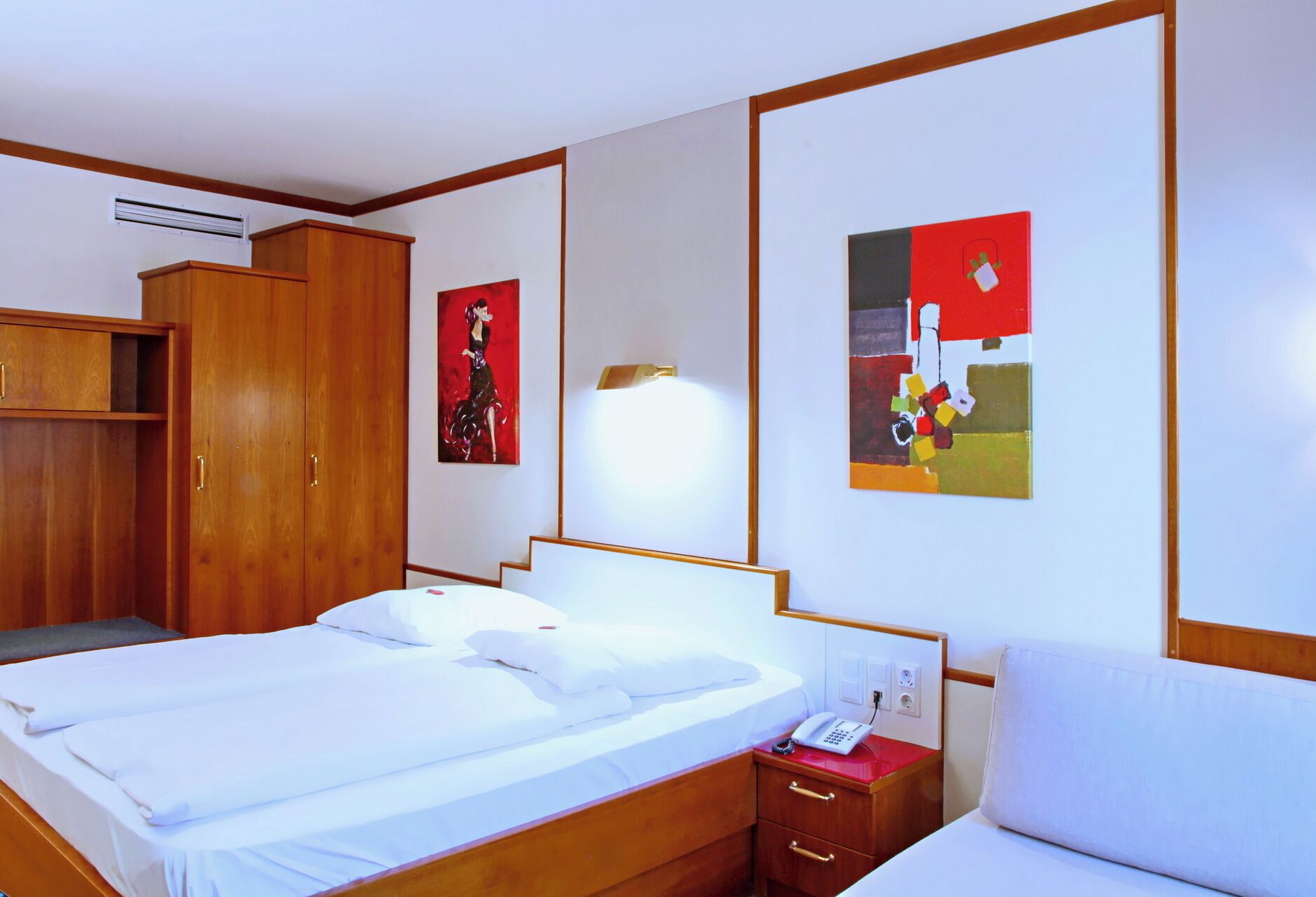 Una habitación de hotel con cama, un armario de madera, una mesita de noche con teléfono y dos cuadros en la pared.