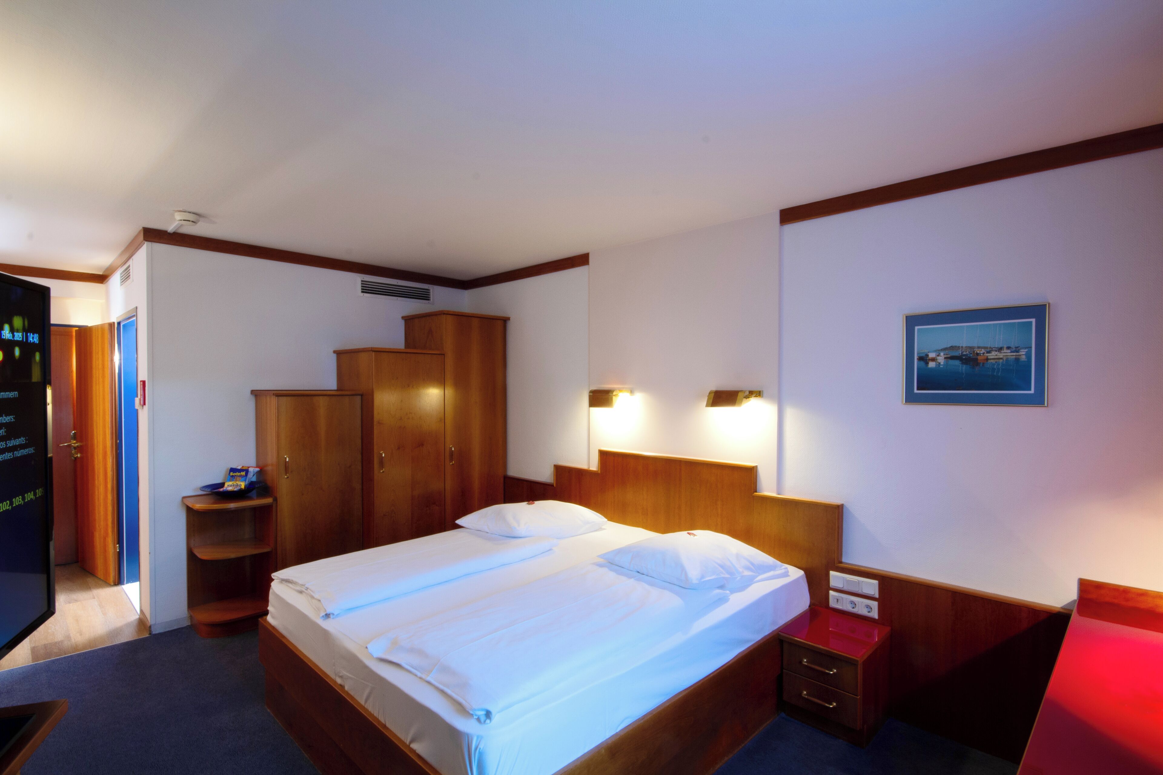 Habitación de hotel con cama, armario de madera, mesita de noche y un cuadro en la pared.