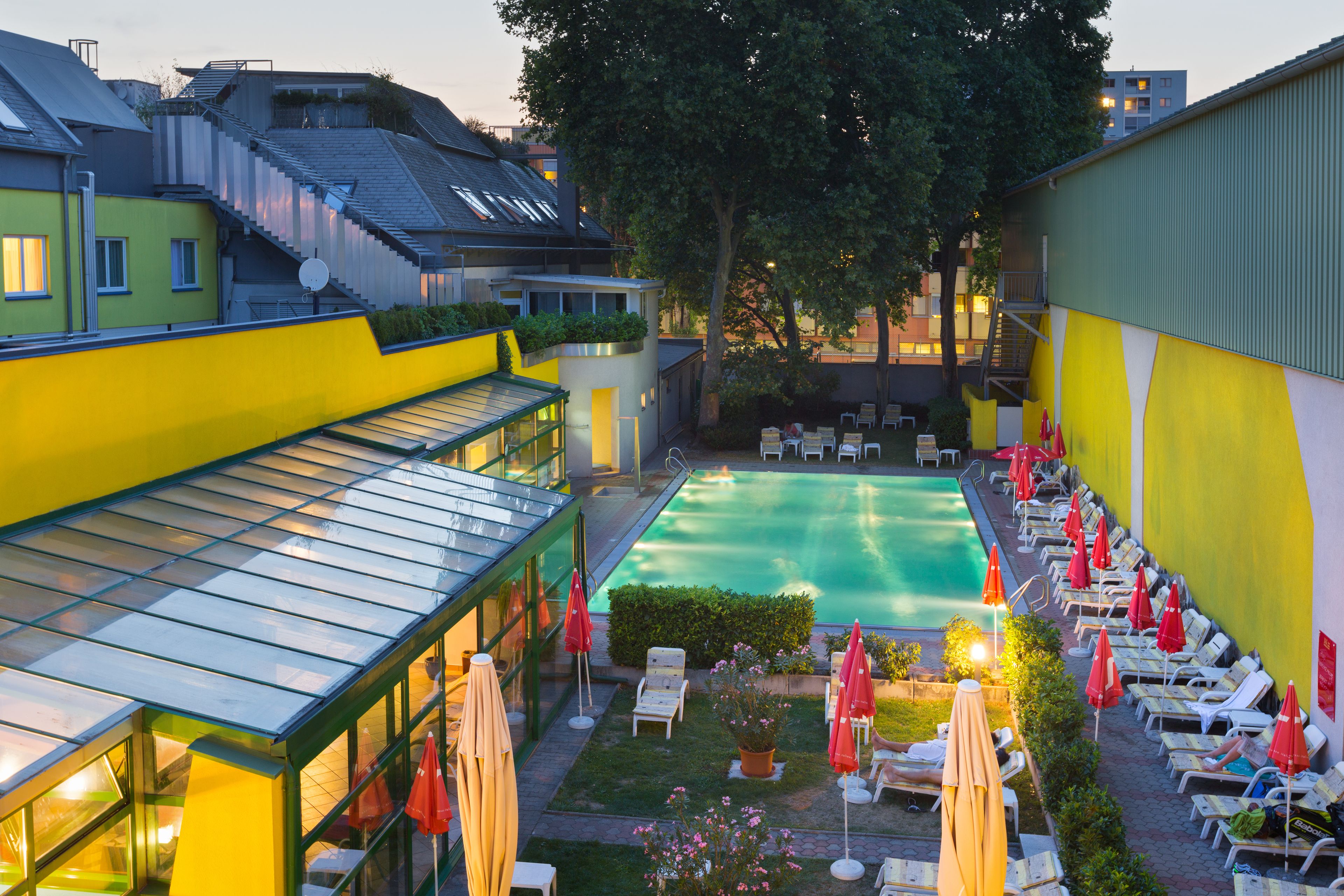 Piscina cercada por espreguiçadeiras e guarda-sóis, com um edifício com cobertura de vidro de um lado.