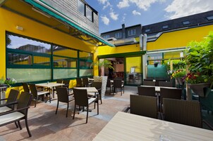 Uma área de pátio externa com mesas, cadeiras, um toldo verde e plantas em vasos.