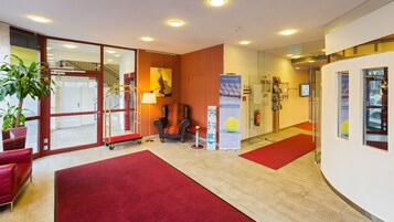 Um saguão de hotel com carpete vermelho, uma área de estar com um sofá vermelho, uma poltrona e uma mesinha com abajur.