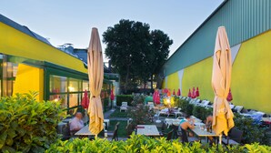 Un área de comedor al aire libre con paredes amarillas, plantas verdes y mesas con sombrillas.