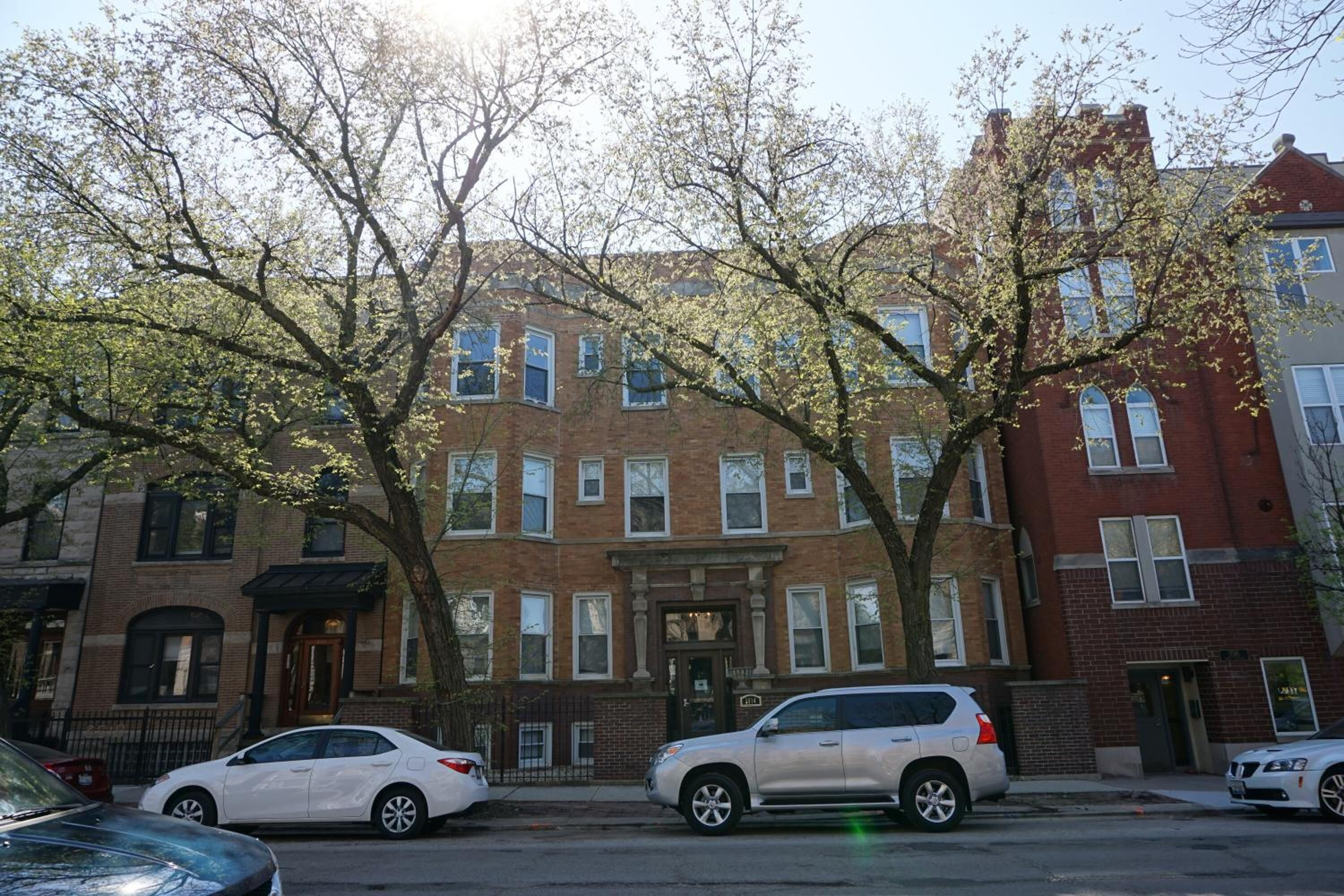 A row of brick townhouses with a central entrance, parked cars, and trees in the foreground.
