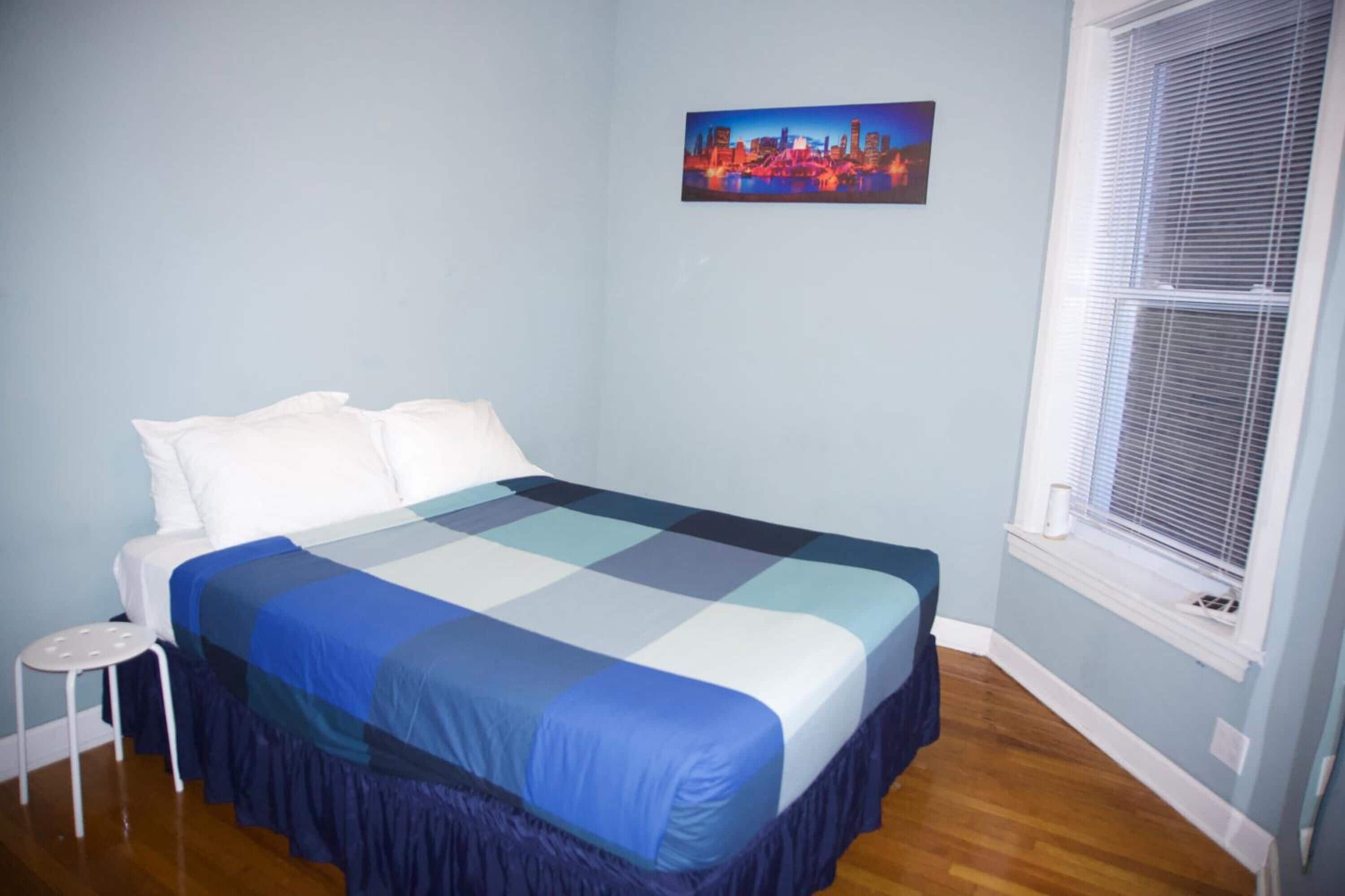 A bedroom with a bed, a small stool, a window with blinds, and a cityscape painting on the wall.