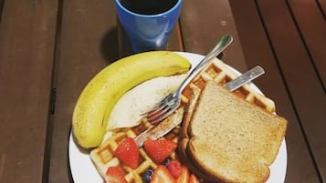 Un plato con waffles, fresas, arándanos, un plátano y tostadas, acompañado de una taza azul y una manzana verde sobre una mesa de madera.