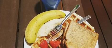 A plate with waffles, strawberries, blueberries, a banana, and toast, accompanied by a blue cup and a green apple on a wooden table.