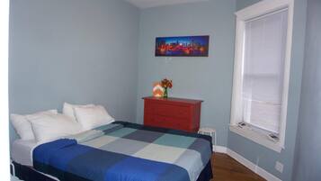 A bedroom with a bed, a nightstand, a window with blinds, and a cityscape painting on the wall.