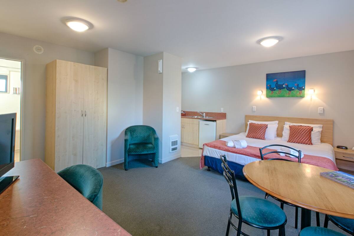 Une chambre d’hôtel comprenant un lit, un bureau, une chaise, une armoire, un coin cuisine et un tableau au mur.