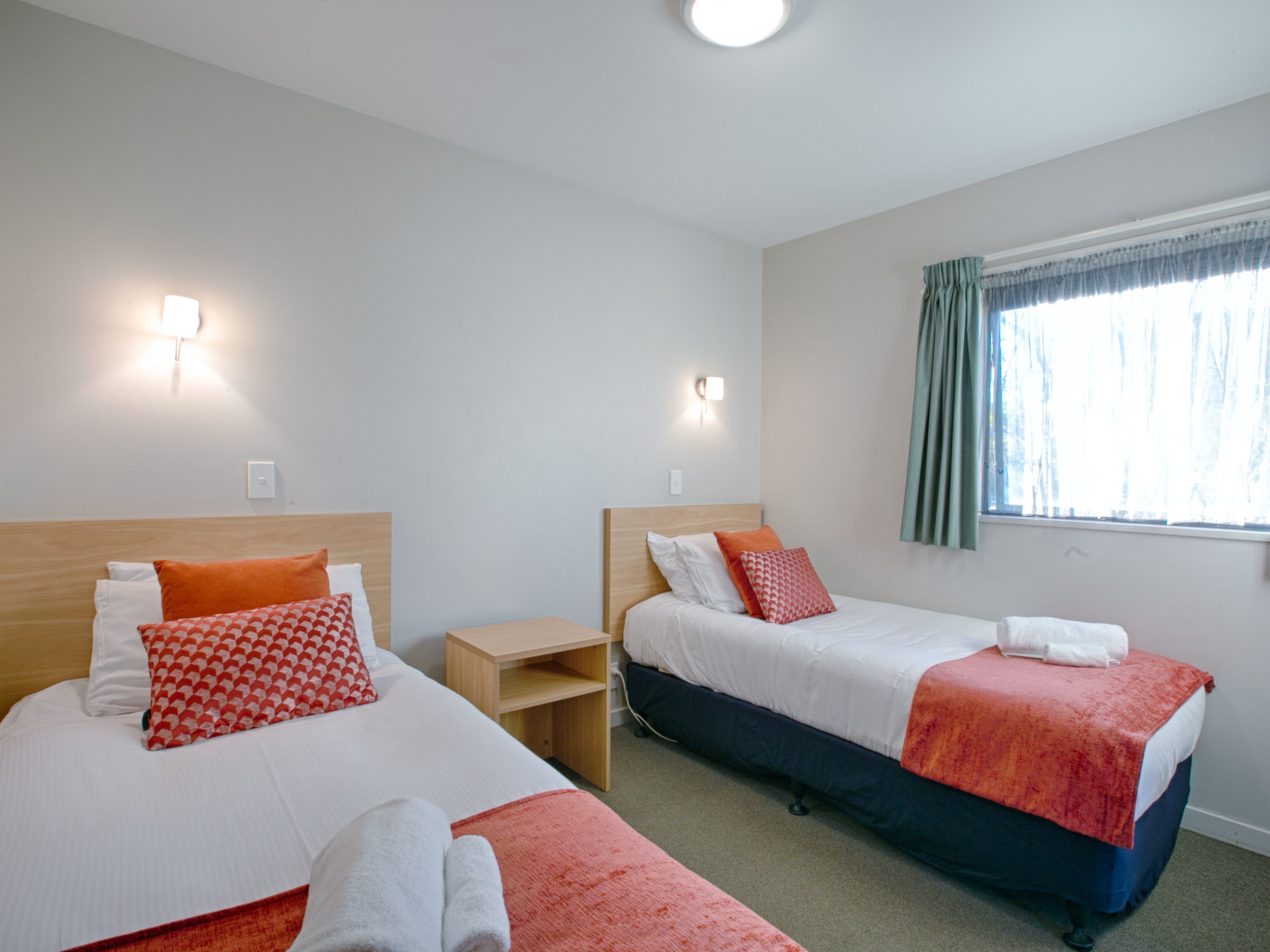 A hotel room with two beds, each with a wooden headboard, white bedding, and red and white patterned pillows. There is a small wooden nightstand between the beds, a window with curtains, and a ceiling light.