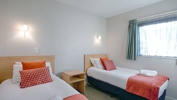 A hotel room with two beds, each with a wooden headboard, white bedding, and red and white patterned pillows. There is a small wooden nightstand between the beds, a window with curtains, and a ceiling light.