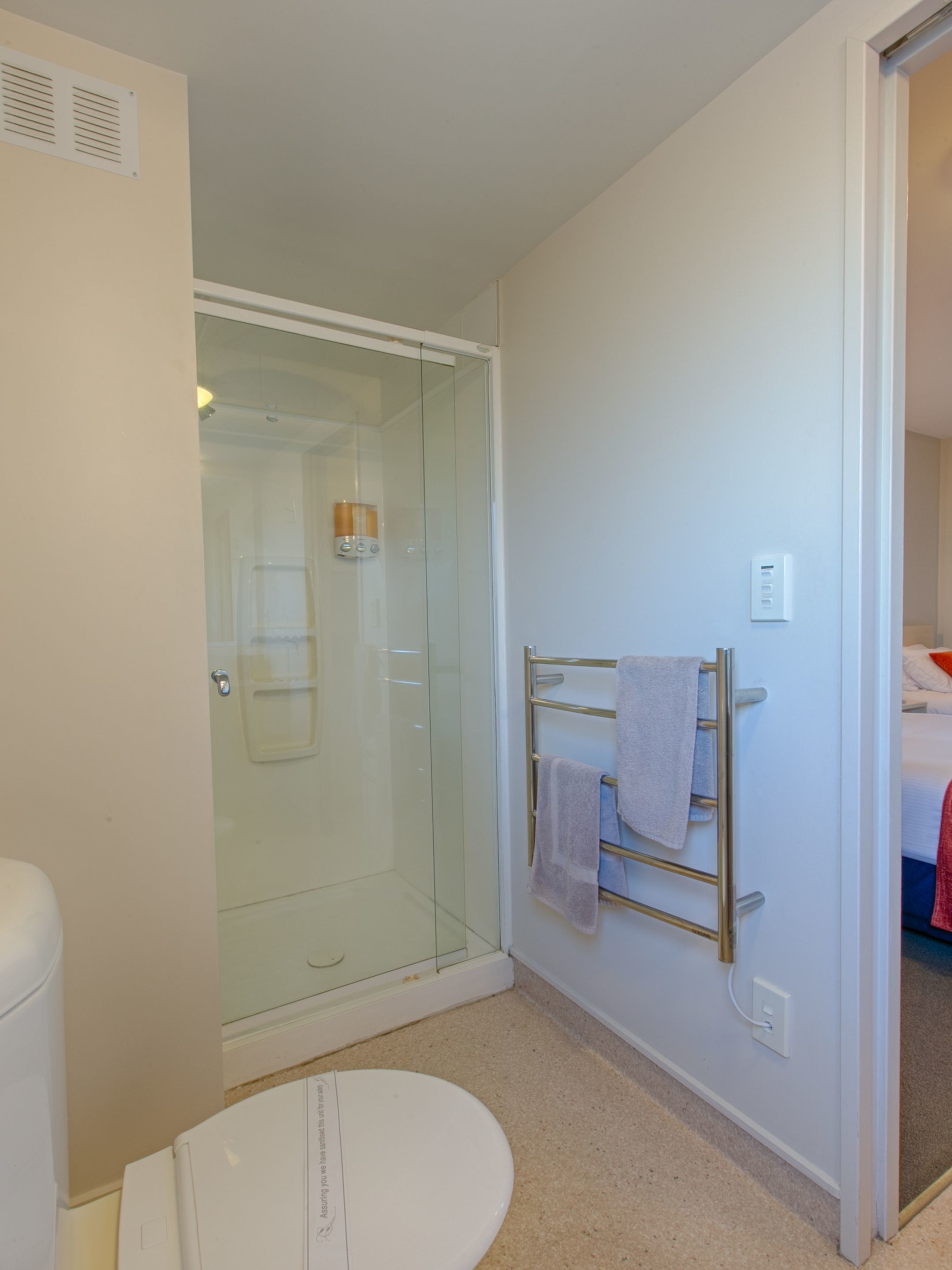 A bathroom with a glass-enclosed shower, a towel rack with towels, a toilet, and a visible electrical outlet.