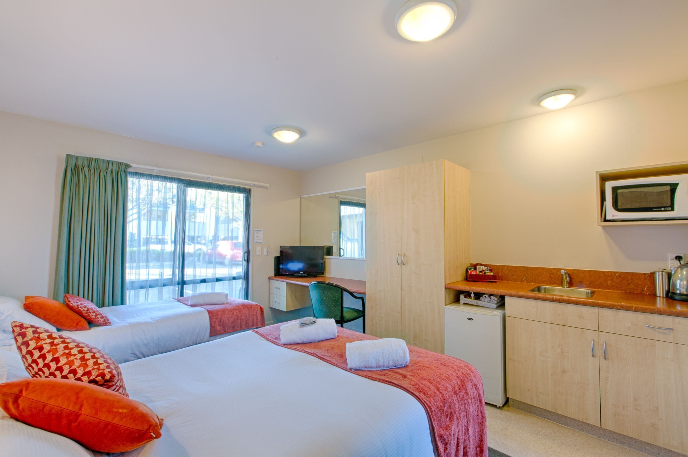 A hotel room with two beds, a desk with a television, a small kitchenette, and a window with curtains.