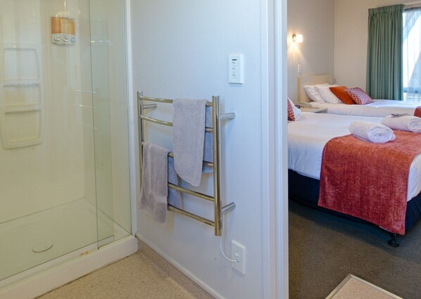 A hotel room with a glass shower enclosure, a towel rack with towels, a bed with a red blanket, and a bedside table with a lamp. - Bella Vista Motel Blenheim (Blenheim)
