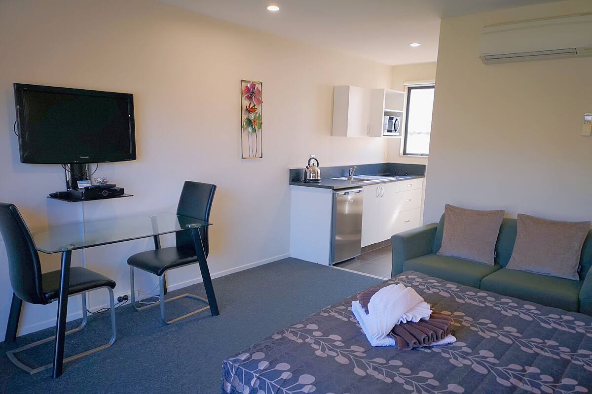 A compact hotel room with a bed, a TV, a small kitchenette, a dining area, and a sofa.