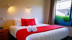 A neatly made bed with white and red bedding, a nightstand with a lamp, and a window with a view of greenery. - Aston Court Motel Blenheim (Blenheim)