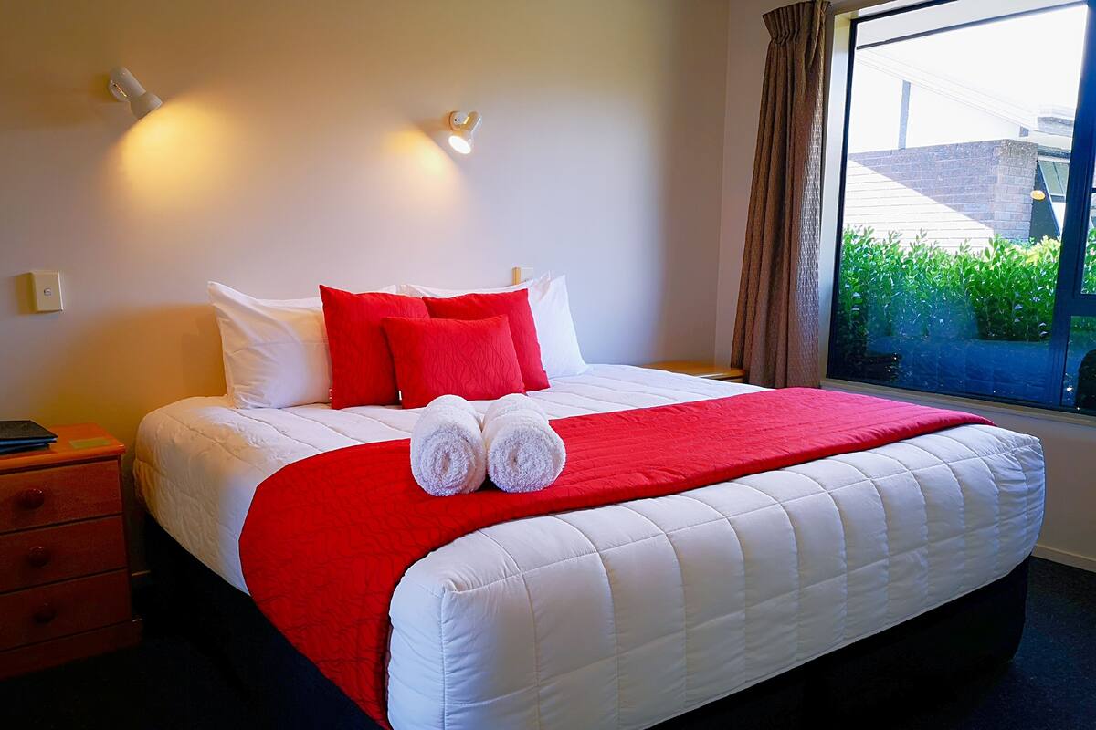 A neatly made bed with white and red bedding, a nightstand with a lamp, and a window with a view of greenery.
