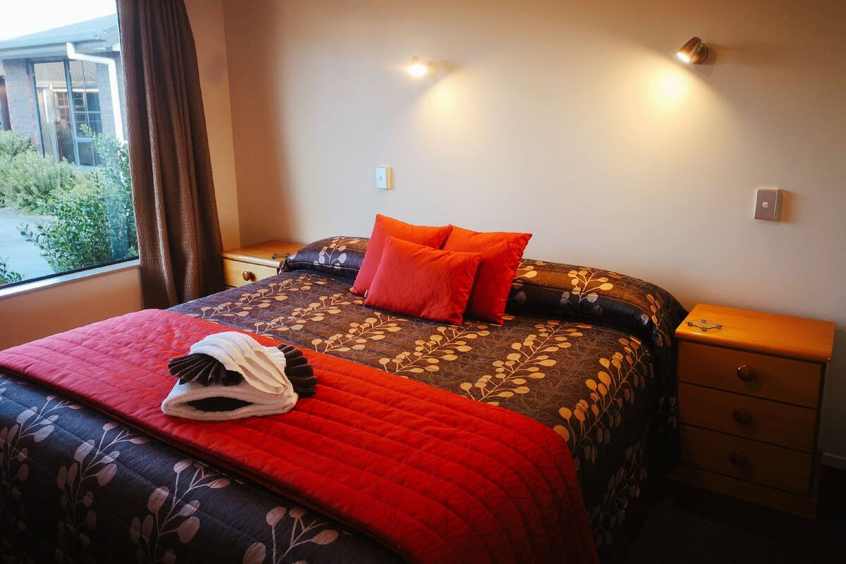 A neatly made bed with a floral-patterned duvet and two bright orange pillows, a wooden nightstand with a drawer, and a window with curtains.