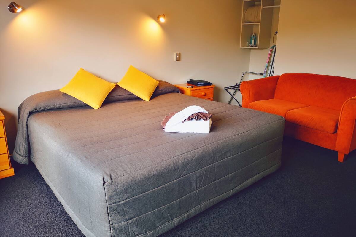 A neatly made bed with a grey bedspread and two yellow pillows, a small orange side table with a book and a lamp, and an orange sofa in a room with a simple, clean design.