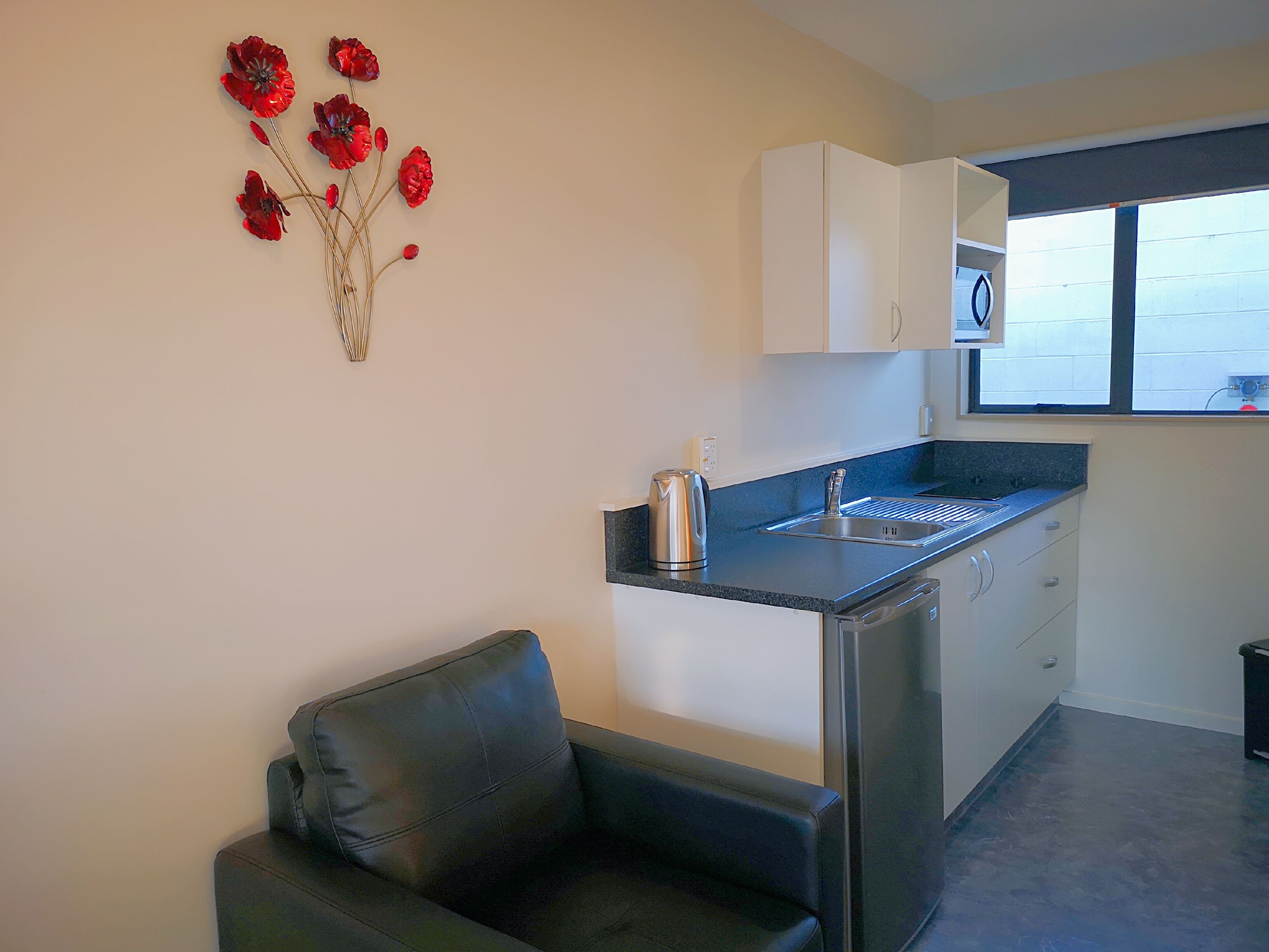 A compact kitchen with a sink, kettle, and cabinets. There's a black leather chair and a decorative red flower arrangement on the wall.