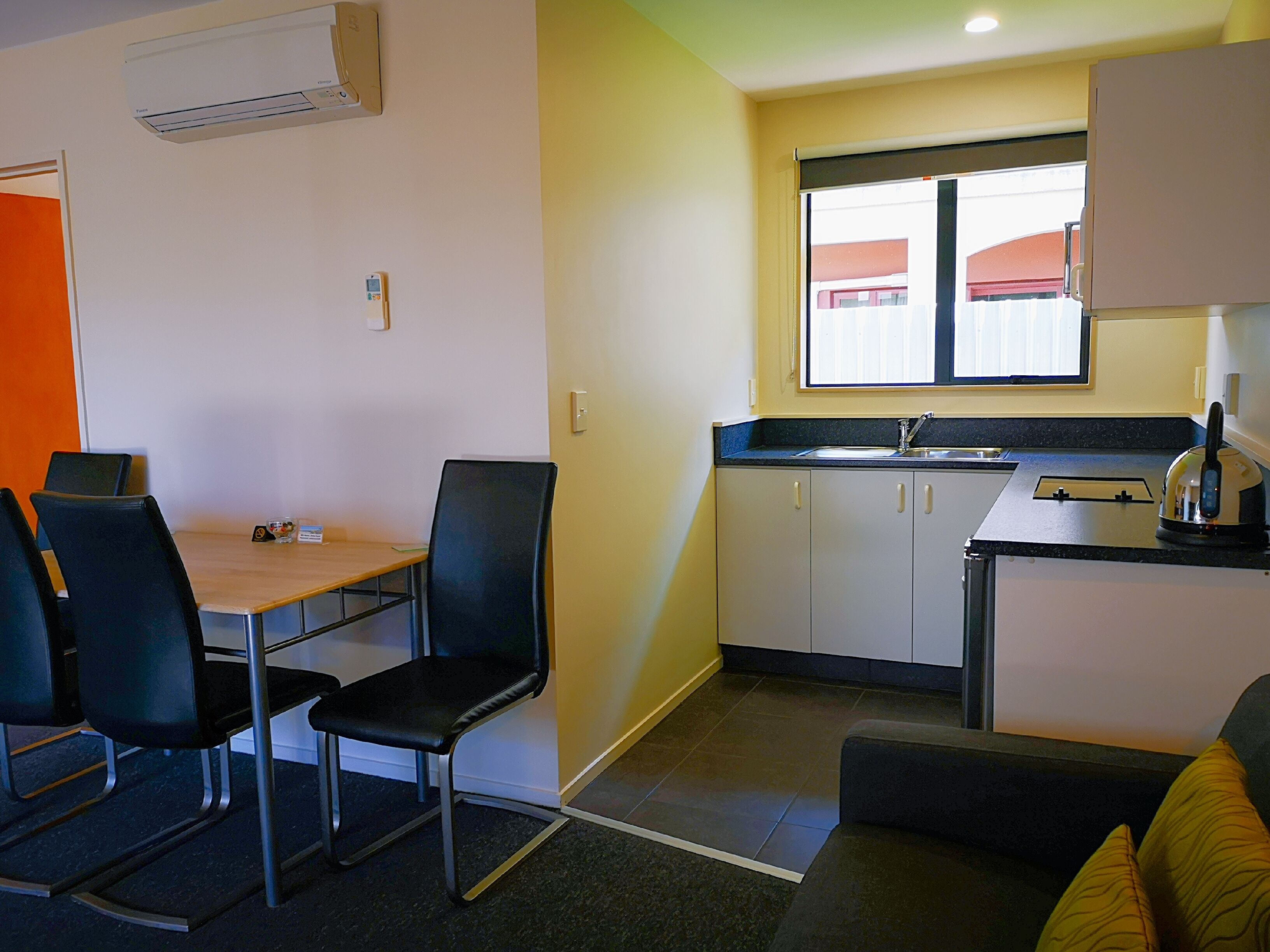 A compact kitchen with a sink, a kettle, and a window. There is a dining area with chairs and a table.