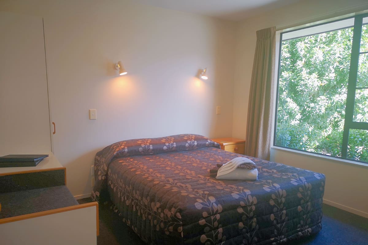 A bedroom with a bed, a nightstand, a window with curtains, and a small desk with a chair.