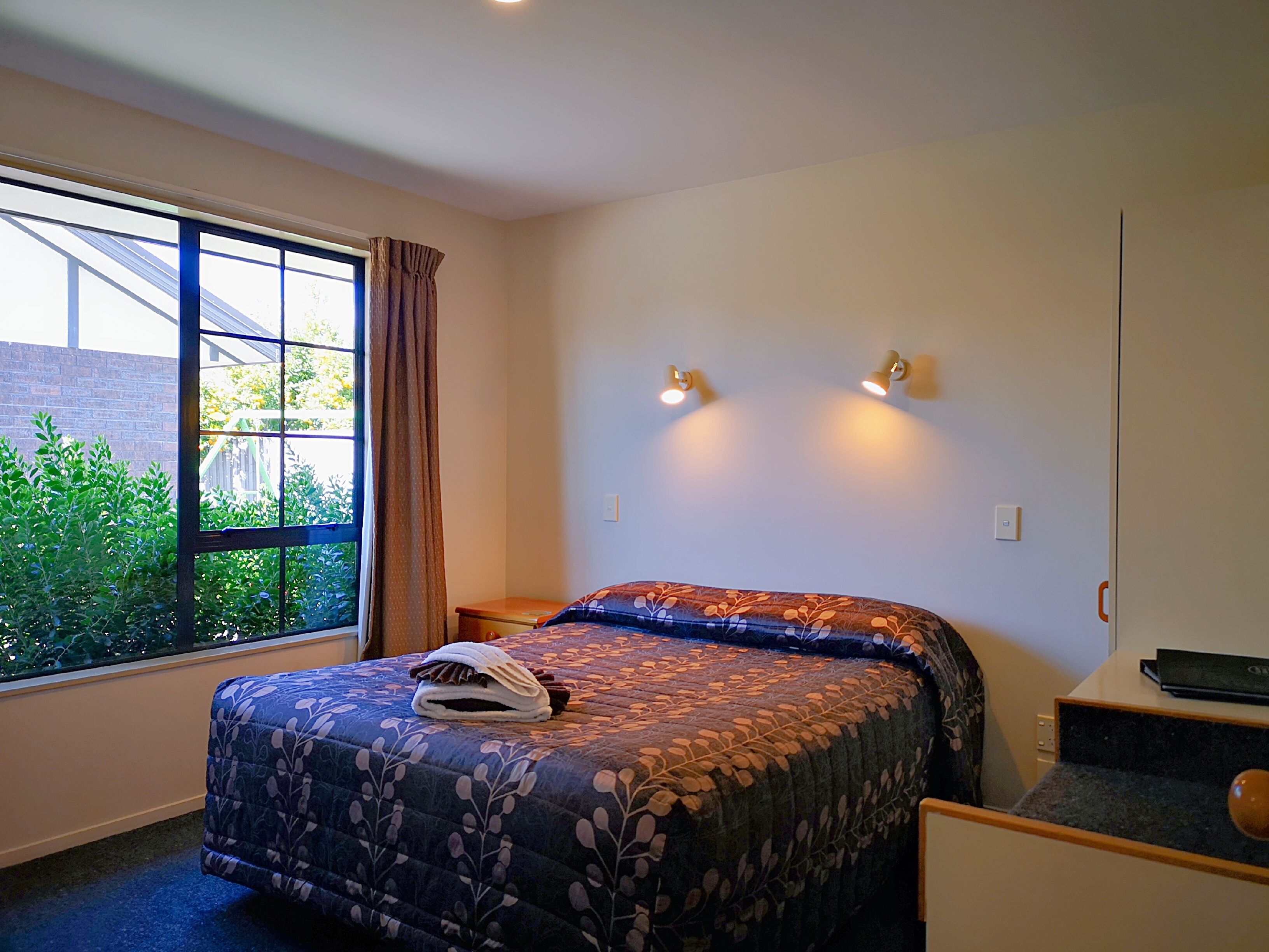 A bedroom with a bed, a window with curtains, a bedside table, and a wall-mounted light fixture.