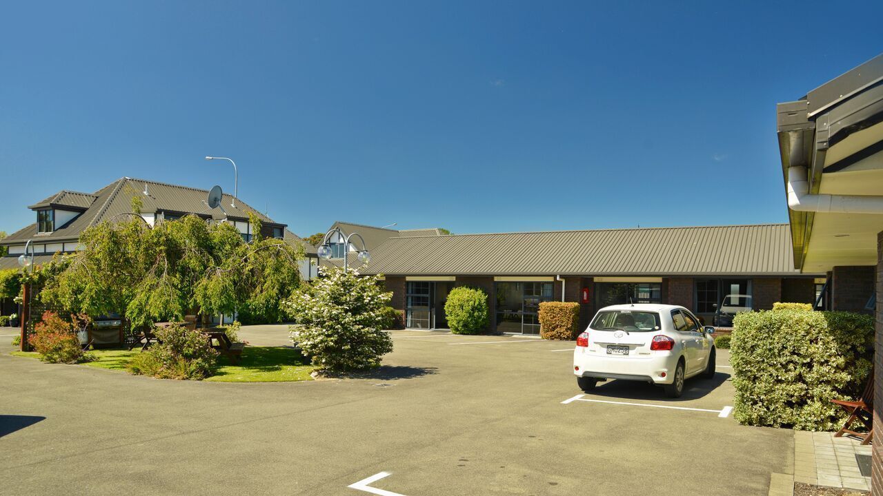 A parking lot with a white car, a building with a flat roof, and some greenery.